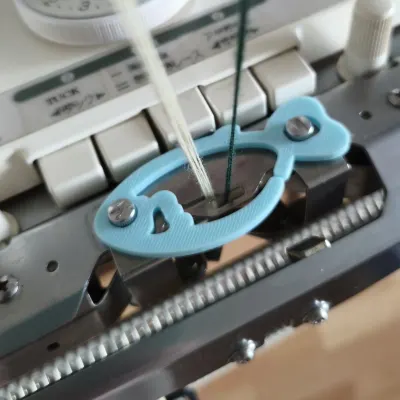 Blue fish-shaped plating yarn holder mounted on a Brother double bed knitting machine